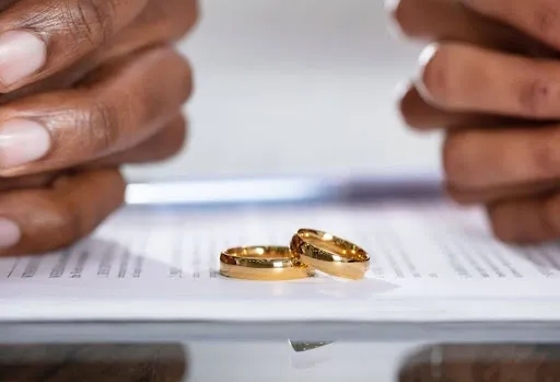 Primer plano de dos anillos de boda de oro en un documento legal, con las manos entrelazadas a ambos lados, lo que sugiere un acuerdo de divorcio o separación.