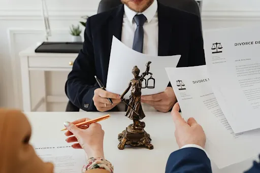 Un abogado con traje revisa los documentos de divorcio con dos clientes. En el centro se encuentra una estatua de bronce de la Dama de la Justicia, que simboliza el juicio legal. El ambiente es formal y serio.